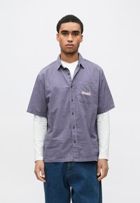 Rövid ujjú, gombos ing ibolyaszínben, mellkasi zsebbel. Finom, függőleges csíkos mintázattal és világos rózsaszín hímzett részletekkel.