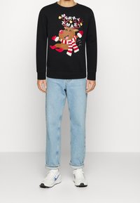 Maglione nero con una renna sorridente che indossa una sciarpa a righe rosse e guanti, jeans azzurri e sneakers bianche con dettagli blu.