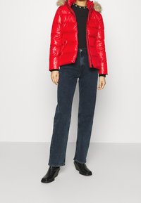 Rød, blanke pufferjakke med pelsbesat hætte, båret over en sort top og parret med mørkeblå jeans og sorte ankelstøvler.