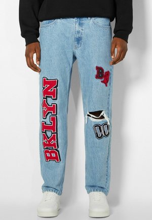 Jean bleu clair avec des patchs rouges et noirs, y compris "BIKINI" sur la jambe gauche et un trou déchiré au genou droit avec un patch "00" en dessous.