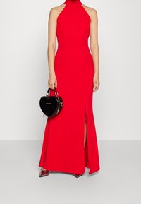 Röd halterneck-klänning med en sidoflik, slät textur och åtsittande design. Matchad med en glansig svart handväska i form av ett hjärta.