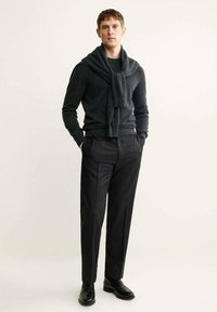 Dunkelgrauer Wollpullover mit einem über die Schulter gehängten Schal, kombiniert mit schwarzen, gestreiften Hosen und schwarzen Slippern.