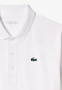 Vit polo-shirt med texturerad tyg, tre knappar och en grön broderad krokodil-logotyp på vänster bröst. Regular fit-design.