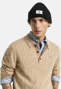 Man in een beige kabeltrui over een lichtblauw overhemd met knopen en een zwarte beanie met opgerolde mouwen en zichtbare tattoos op de onderarmen.