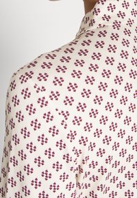 Camicia a maniche lunghe in tessuto color crema con un piccolo motivo geometrico ripetuto in bordeaux e viola. Presenta un colletto strutturato e una superficie liscia.