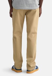 Beige chinos made of cotton with a straight leg design. Visible stitching and side pockets. Paired with blue and white sneakers.