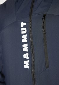 Navyblauwe technische jas met een ritszak en een groot wit "MAMMUT" logo, gemaakt van textuurstof en met een contrasterende zwarte rits.