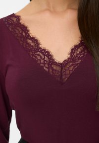 Gros plan sur un haut bordeaux avec un décolleté en V bordé de dentelle délicate et les longs cheveux noirs d'une femme sur un côté.