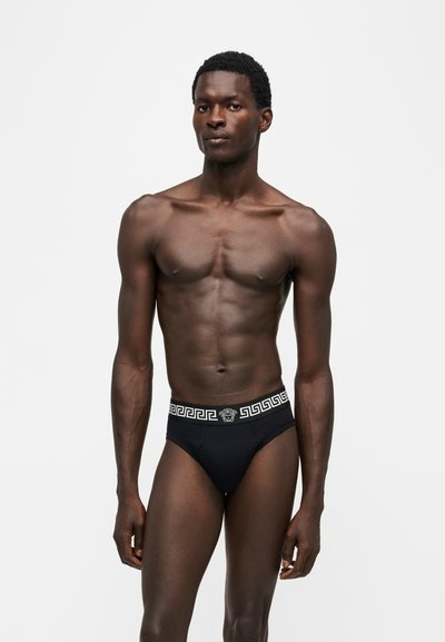 Homme musclé à la peau foncée debout en slip noir avec motif clé grecque et logo Méduse sur la ceinture, sur fond blanc uni.