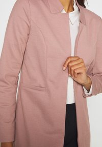 Blazer rosa chiaro con un design senza colletto, tessuto testurizzato e una tasca frontale. Il blazer è abbinato a una camicia bianca sottostante.