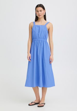 Nő ujjatlan, térdig érő kék ruhában, összegyűrt derékrésszel és négyzet alakú nyakkivágással, mezítláb áll egy egyszínű világos háttér előtt.