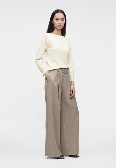 Femme aux longs cheveux foncés debout, vêtue d'un haut crème à manches longues et d'un pantalon large beige avec une ceinture, les mains partiellement dans les poches.