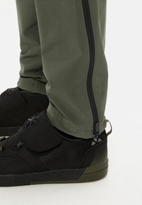 Groene sportbroek met een zijrits, gecombineerd met zwarte mesh en rubberen sneakers met groene accenten en een strap-sluiting.