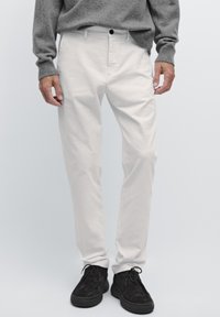 Weiße Baumwollhosen in Slim Fit mit seitlichen Taschen und einer vorderen Knopfleiste, kombiniert mit schwarzen Wildleder-Schnürschuhen.