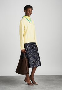 Žltý sveter s vysokým golierom, modré a zelené akcenty, kvetinová sukňa so námorníckymi flitrami, hnedá semišová taška a béžové vysoké podpätky.