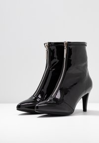 Par de botines de charol negro con cierres plateados en la parte frontal y tacones de altura media sobre una superficie blanca.