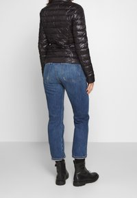 Svart vadderad jacka med en glänsande yta, kombinerad med blå denimjeans. Jackan har en figursydd design och hög krage. Svarta ankelskor fulländar looken.