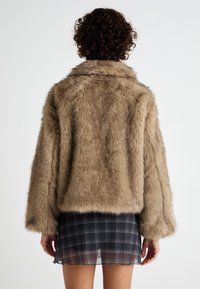 Brun kunstpelsjakke med krage, vide ermer og kort passform, brukt over et svart rutete mini skjørt.