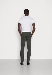 Mężczyzna stoi przodem do białej ściany, ubrany w białą koszulkę z krótkim rękawem, ciemnoszare spodnie i czarne buty wizytowe, z rękami w kieszeniach.