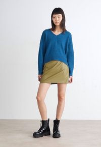 Modra textured oversize pulover s globokim V-izrezom, v kombinaciji s prilegajočo olivno zeleno usnjeno mini krilo in debelimi črnimi vezalkami.
