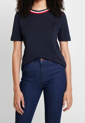 Vrouw draagt een marineblauw aansluitend T-shirt met een rood-wit-blauw gestreepte kraag, gecombineerd met een hoge taille donkere blauwe spijkerbroek.