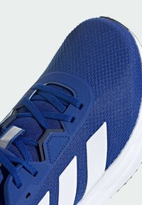 Modrá atletická bota s texturovaným síťovaným svrškem, vybavená bílými pruhy a kontrastními tkaničkami. Obsahuje zesílenou patu a elegantní design.