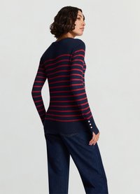 Navy striped sweater with red horizontal lines, fitted design, ribbed cuffs, and three-button accents on sleeves, paired with blue denim pants.