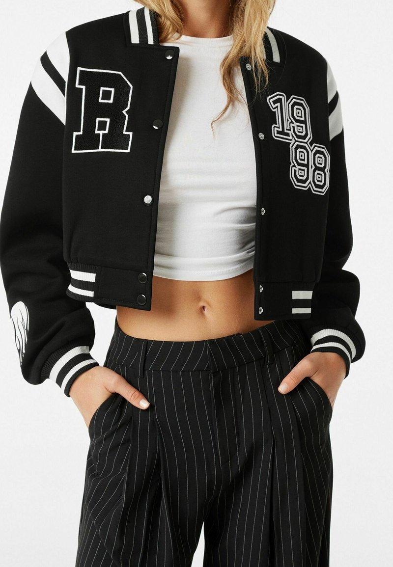 Black cropped varsity jacket with white striped sleeves, featuring a letter "R," embroidered numbers "1998," and a smooth texture. Paired with pinstriped pants.
