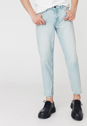 Jeans en denim bleu clair au style droit, présentant une texture légèrement délavée et des poignets effilochés, associés à des chaussures noires à lacets.