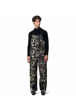 Giovane uomo che indossa una maglia nera a maniche lunghe e pantaloni con bretelle mimetici con caviglie rinforzate nere, in piedi che guarda verso destra.