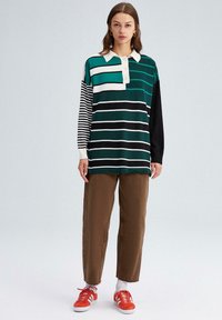 Garais piedurknes polo krekls ar zaļām, melnām un baltām svītrām; melni un balti svītroti piedurknes; ar četru pogu aizdari; kombinēts ar brūniem biksēm un sarkaniem sporta apaviem.