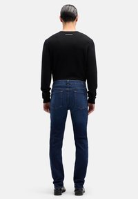 Mężczyzna stojący tyłem, ubrany w czarną koszulę z długim rękawem, ciemnoniebieskie dżinsy i czarne buty, na jednolitym białym tle.