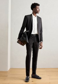 Costum negru, croit pe măsură, cu o cămașă albă, curea neagră și mocasini. Ținând o geantă mare din piele neagră. Design slim fit, textură de țesătură netedă.