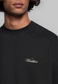 Svart sweatshirt i texturerat tyg med rund hals och en broderad logotyp i vitt på vänster bröst.