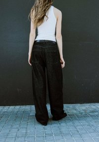 Czarne dżinsy o szerokich nogawkach z teksturowanej tkaniny, z wysokim stanem, dwoma tylnymi kieszeniami i paskiem. Modelka nosi biały top na ramiączkach.