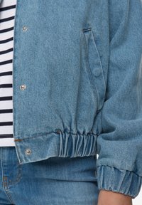 Světlomodrá džínová bunda s elastickým pasem a manžetami, s přezkami na patentky a pohodlným střihem. Pod ní je nošena proužkovaná košile.