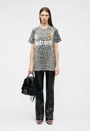 Vrouw draagt een oversized grijs t-shirt met luipaardprint en de tekst "just cavalli", zwarte leren broek, sandalen en houdt een zwarte rugzak vast.