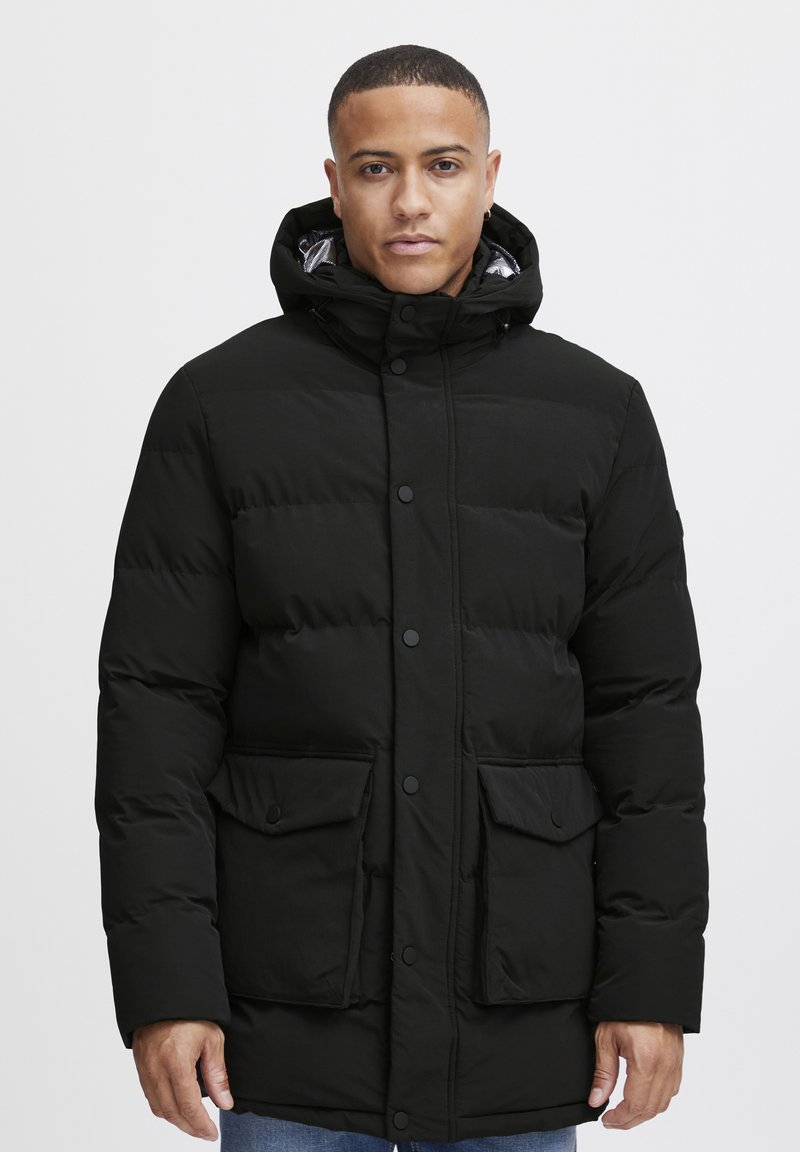 Chaqueta de plumas negra con capucha. Presenta un cierre de botones al frente, dos bolsillos frontales y una textura acolchada. Diseño casual adecuado para el invierno.