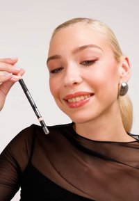 Svart sminkpenna med ett klart lock, hålls av en person i en genomskinlig svart topp. Pennan har en krämig, ljus spets. Mjuk finish.