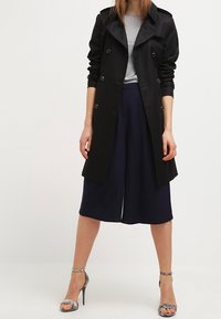 Abrigo negro de trench de tela suave, diseño de doble botonadura, combinado con culottes azul marino y sandalias de tacón metálico.