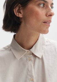 Light beige collared shirt with a smooth texture, featuring a button placket and simple, classic design. Buttons are light and round.