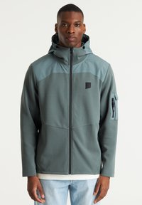 Donkergroene hooded jas met lichtblauwe schouderpanelen. Voorzien van ritssluiting en zijzakken. Gladde stof, relaxte pasvorm.