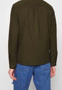 Oliwkowa koszula z długimi rękawami, mankietami na guziki i detalem yoke na plecach. Tkanina wydaje się miękka; stylizowana z niebieskimi jeansami.
