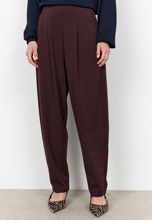 Person wearing dark brown high-waist pleated trousers and zebra-print pointed-toe heels, standing against a plain white wall.