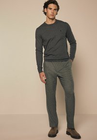 Grauer, gestrickter Pullover mit Rundhalsausschnitt, kombiniert mit grünen karierten Hosen. Das Model trägt braune Wildlederschuhe, die eine maßgeschneiderte Passform und Textur zeigen.