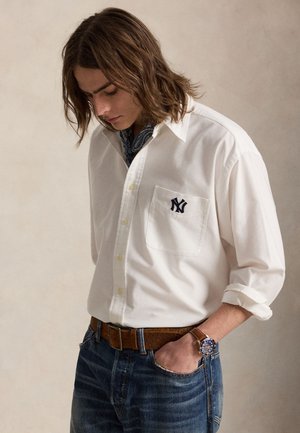 Νεαρός άνδρας με μακριά μαλλιά μέχρι τον ώμο, φοράει λευκό πουκάμισο με το λογότυπο NY, μπλε τζιν, καφέ ζώνη, μπλε ρολόι και ένα φουλάρι γύρω από το λαιμό του.