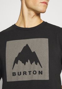 Černé bavlněné tričko s šedým čtvercovým grafikou, která zobrazuje siluetu hory a slovo "BURTON" tučně napsané pod ní.