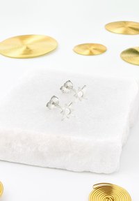 Orecchini a perno in argento a forma di stella con un centro circolare, posizionati su una superficie di pietra bianca, circondati da accenti circolari dorati.