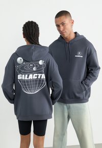 Felpa con cappuccio grigia scura caratterizzata da una grafica planetaria sul retro con il testo "GALATTICO". Sul davanti c'è un piccolo logo con la scritta "DIVERTIMENTO COSMICO". Materiale in misto cotone.