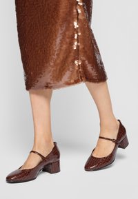 Brown patent leather Mary Jane heels with a crocodile texture, square toe, and chunky heel. Accompanied by a shimmery bronze sequin skirt.